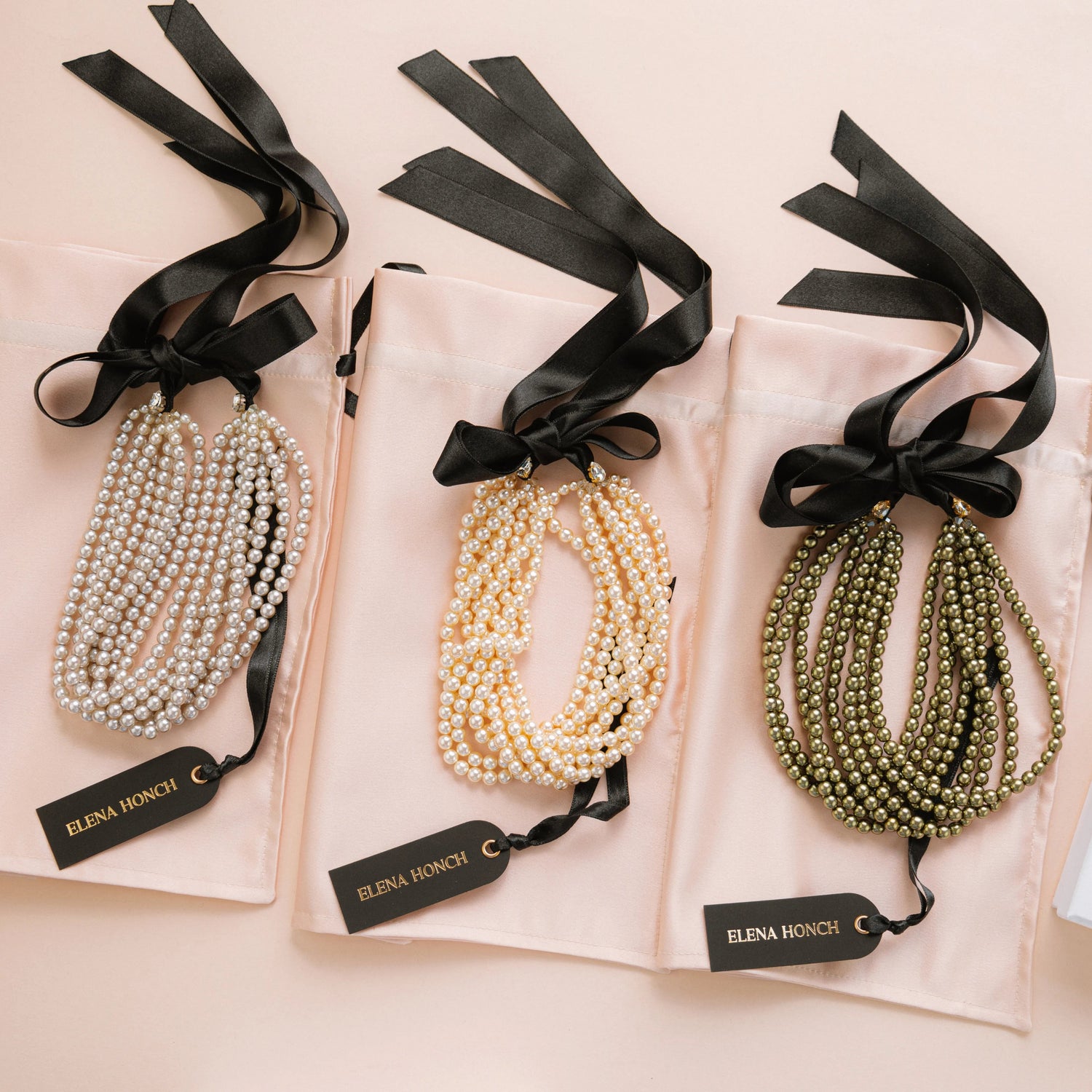 Three strands of multi-layered beaded necklaces in white, cream, and olive green, each presented on a light pink satin pouch with a black ribbon and tag. A white Elena Honch box is visible on the right.